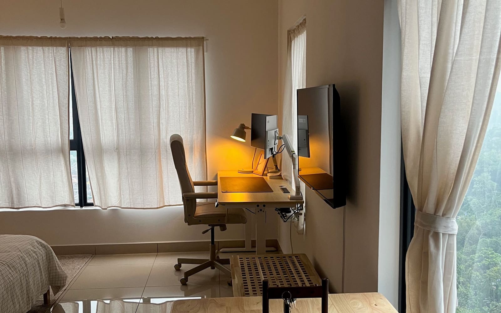 Bright studio with a standing desk, dual monitors and a warm lamp beside linen-curtained windows